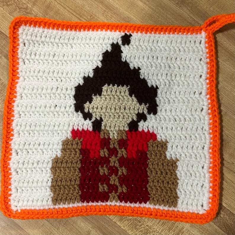 May include: A crocheted square with an orange border featuring a pixelated image of a person with brown hair wearing a red and brown outfit.