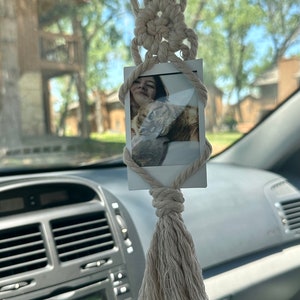 Op de afbeelding: Een witte macramé autohanger met een kwast die aan de achteruitkijkspiegel van een auto hangt. Een wit fotolijstje met een foto van een persoon en een kat is aan de macramé bevestigd.