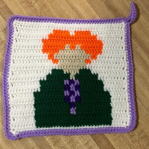 May include: Crocheted square with a purple border and a cartoon image of a person with orange hair, a green shirt, and a purple flower.