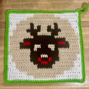 May include: A square, crocheted potholder featuring a pixelated design of a black creature's face with white teeth and red accents. The potholder has a green border and a loop for hanging. The background is tan, white, and brown.