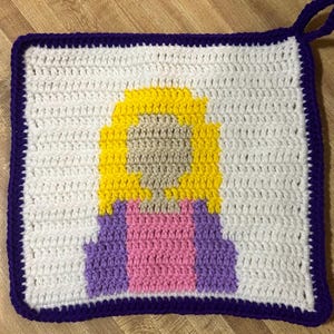 May include: Crocheted square hot pad with a purple border and a pixelated image of a person with yellow hair, a pink shirt, and a purple jacket.