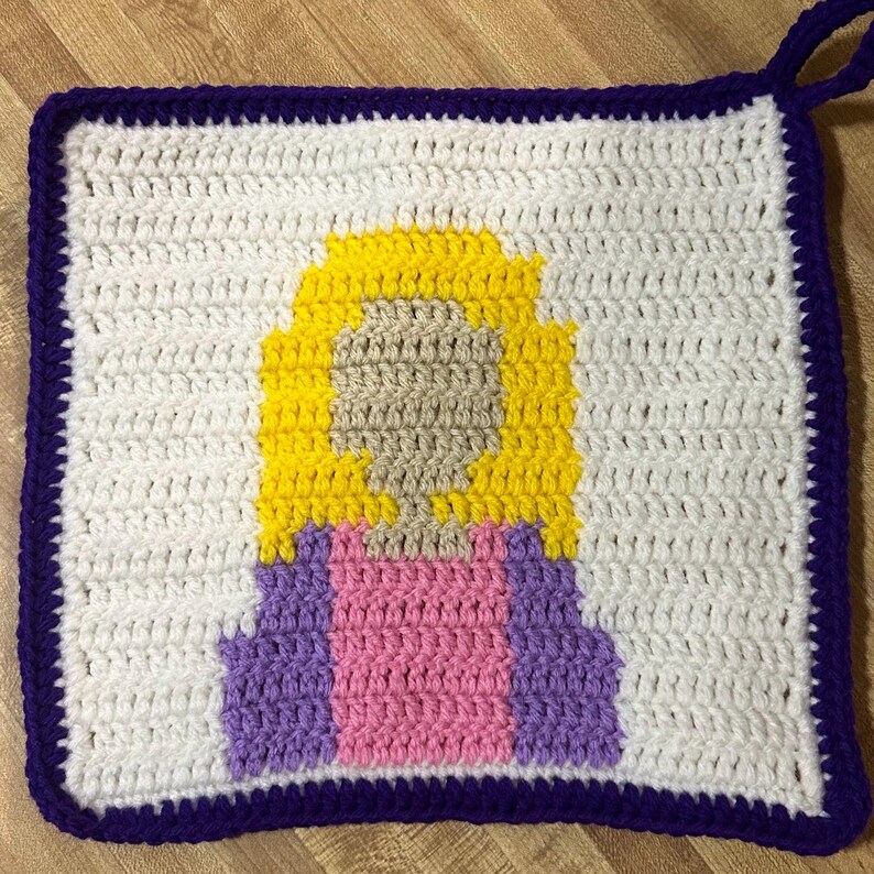 May include: Crocheted square hot pad with a purple border and a pixelated image of a person with yellow hair, a pink shirt, and a purple jacket.
