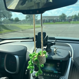 May include: A crocheted plant decoration hanging from a car's rearview mirror. The plant has a small, beige pot and green, string-like leaves. The car's interior includes a dashboard, radio, and a phone holder.