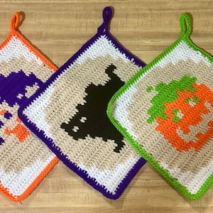 Könnte beinhalten: Drei gehäkelte Topflappen mit Halloween-Motiven. Der erste Topflappen ist lila und weiß mit einem Geistermotiv. Der zweite Topflappen ist schwarz und weiß mit einem Fledermausmotiv. Der dritte Topflappen ist orange, grün und weiß mit einem Kürbismotiv.