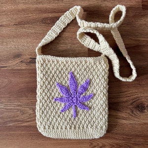 May include: A crocheted shoulder bag with a purple marijuana leaf design. The bag is made of a beige yarn and has a long strap.