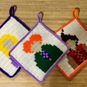 May include: Three crocheted pot holders featuring different characters. The first pot holder is purple and features a yellow character. The second pot holder is lavender and features an orange character with green and purple clothing. The third pot holder is orange and features a brown character with red and brown clothing.
