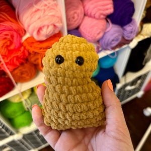 May include: A crocheted plush toy shaped like a person with a brown body and black eyes. The toy is being held in a hand.
