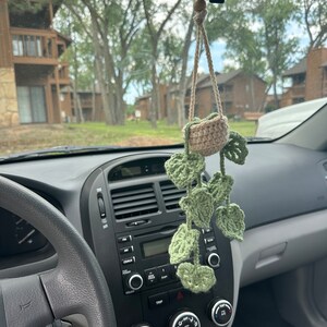 May include: A crocheted plant hanger with a small pot and green leaves hanging from the rear view mirror of a car.