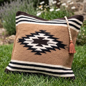 K&ouml;nnte beinhalten: Ein quadratisches Zierkissen mit einem geometrischen Muster in Braun, Schwarz und Wei&szlig;. Das Kissen hat eine Quaste und Perlendetails. Das Kissen liegt auf gr&uuml;nem Gras.