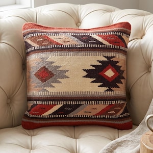 May include: A square throw pillow featuring a woven geometric pattern in shades of rust orange, cream, dark brown, and grey. The design consists of horizontal bands with diamond and stepped motifs, creating a textured appearance on the fabric surface.