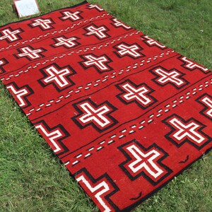 May include: A rectangular woven rug featuring a vibrant red background with a repeating pattern of white crosses outlined in black. The design includes horizontal rows of these crosses separated by thin, dashed black lines across the entire surface.