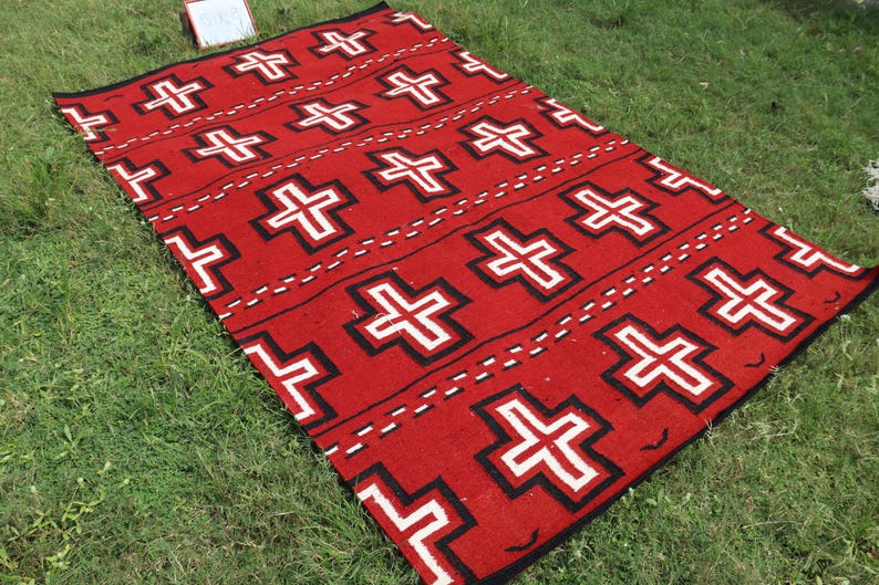 May include: A rectangular woven rug featuring a vibrant red background with a repeating pattern of white crosses outlined in black. The design includes horizontal rows of these crosses separated by thin, dashed black lines across the entire surface.