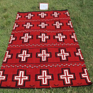 May include: A rectangular woven rug featuring a vibrant red background with a repeating pattern of white crosses outlined in black. The design includes horizontal rows of these crosses separated by thin, dashed white lines across the surface.