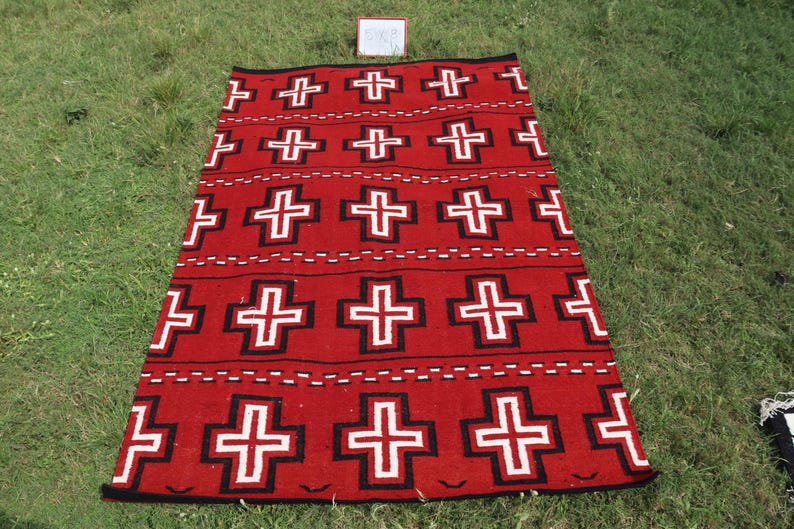 May include: A rectangular woven rug featuring a vibrant red background with a repeating pattern of white crosses outlined in black. The design includes horizontal rows of these crosses separated by thin, dashed white lines across the surface.
