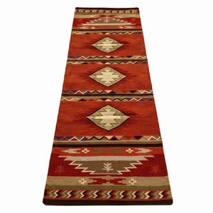 Puede incluir: Una alfombra larga y rectangular con un diseño del suroeste. La alfombra presenta un fondo de color óxido con patrones geométricos en tonos beige, marrón, verde y azul. El diseño incluye formas de diamante y líneas en zigzag.