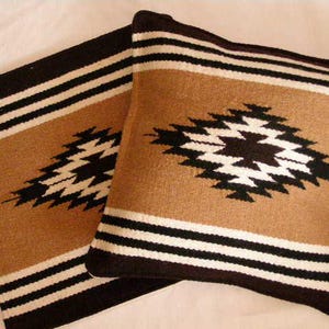K&ouml;nnte beinhalten: Zwei quadratische Kissen mit einem s&uuml;dwestlichen Design. Die Kissen haben einen beigefarbenen Hintergrund mit einem schwarz-wei&szlig;en geometrischen Muster und schwarzen R&auml;ndern. Sie bestehen aus einem gewebten Stoff.
