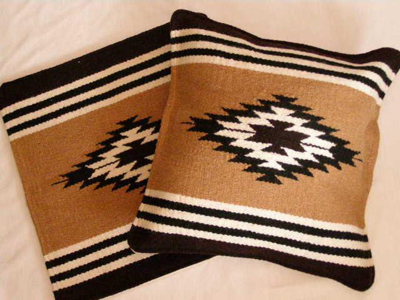 K&ouml;nnte beinhalten: Zwei quadratische Kissen mit einem s&uuml;dwestlichen Design. Die Kissen haben einen beigefarbenen Hintergrund mit einem schwarz-wei&szlig;en geometrischen Muster und schwarzen R&auml;ndern. Sie bestehen aus einem gewebten Stoff.