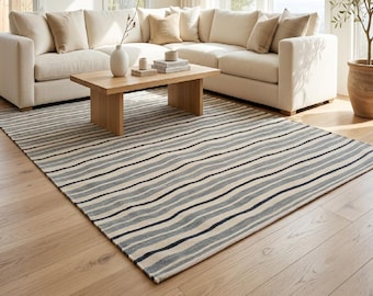 Op de afbeelding: Een rechthoekig vloerkleed met een patroon van horizontale golvende strepen in tinten donkerblauw, lichtgrijs en crème. Het kleed ligt plat op een lichte houten vloer en bedekt een groot deel van de ruimte voor een bank.