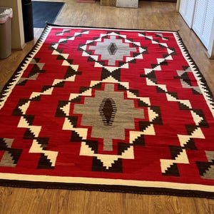 Op de afbeelding: Een rood, wit en bruin patroon tapijt met een geometrisch ontwerp. Het tapijt heeft een franje rand.