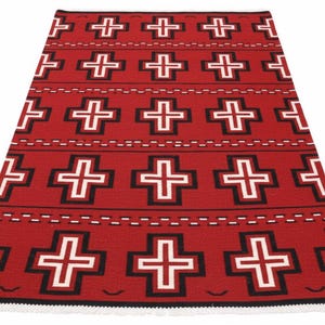 May include: A rectangular woven rug featuring a vibrant red background with a repeating pattern of white crosses outlined in black. The design includes horizontal rows of dashed lines and small curved accents, finished with a white fringe at the edge.