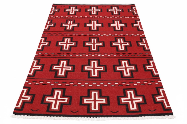 May include: A rectangular woven rug featuring a vibrant red background with a repeating pattern of white crosses outlined in black. The design includes horizontal rows of dashed lines and small curved accents, finished with a white fringe at the edge.