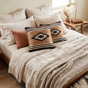 K&ouml;nnte beinhalten: Ein Bett mit neutralfarbener Bettw&auml;sche, darunter Kissen in Creme-, Rost- und Braunt&ouml;nen. Zwei Kissen weisen ein geometrisches Design auf. Das Bett ist mit einer strukturierten Decke und einem gewebten Teppich ausgestattet.