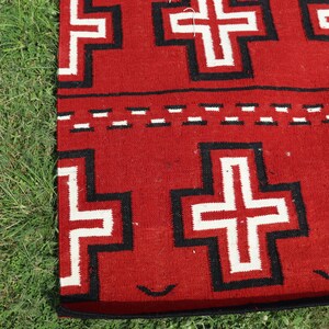 May include: A woven textile featuring a vibrant red background with a repeating pattern of white crosses outlined in thick black borders. A horizontal band with a dashed white and black line separates the rows of crosses on the fabric surface.
