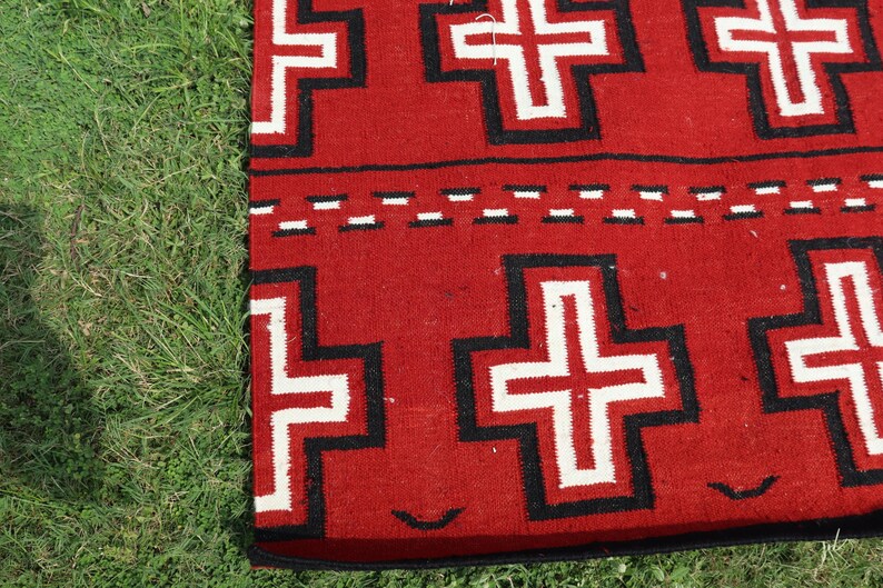 May include: A woven textile featuring a vibrant red background with a repeating pattern of white crosses outlined in thick black borders. A horizontal band with a dashed white and black line separates the rows of crosses on the fabric surface.
