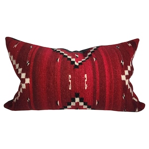 May include: A rectangular decorative pillow with a red background and a woven pattern. The pattern includes vertical stripes, white and black geometric shapes, and a central design. The pillow is designed for home decor.