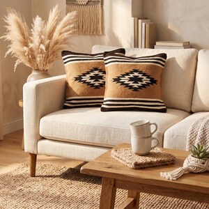 K&ouml;nnte beinhalten: Zwei dekorative Kissen mit einem geometrischen Muster in Schwarz, Beige und Wei&szlig; liegen auf einem cremefarbenen Sofa. Ein Holztisch hat gestapelte Tassen, einen gewebten Untersetzer und eine kleine Pflanze. Der Raum ist in neutralen T&ouml;nen gehalten.