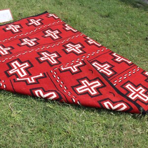 May include: A rectangular woven rug featuring a vibrant red background with a repeating pattern of white crosses outlined in black. The design includes horizontal rows of these geometric cross motifs separated by dashed black lines across the surface.
