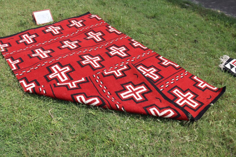 May include: A rectangular woven rug featuring a vibrant red background with a repeating pattern of white crosses outlined in black. The design includes horizontal rows of these geometric cross motifs separated by dashed black lines across the surface.