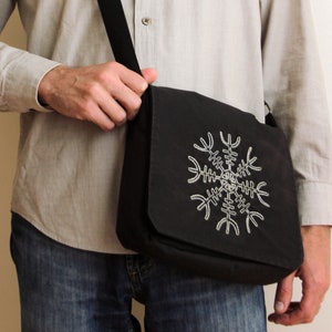 Może przedstawiać: Mężczyzna w jasnobeżowej koszuli z guzikami i niebieskich dżinsach nosi czarną torbę listonoszkę z haftowanym białym wzorem śnieżynki.