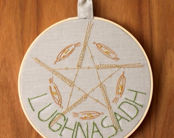 以下が含まれることがあります： 小麦の穂と緑色の糸で「Lughnasadh」という言葉が刺繍された、五芒星模様の刺繍枠アートです。