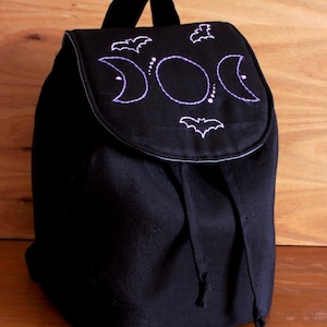 以下が含まれることがあります： 3つの月とコウモリの紫色の刺繍が施された黒いキャンバスのバックパック。