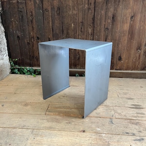 May include: A small, square, metal side table with a light gray finish. The table has a simple, minimalist design with an open base. The table is made of a single piece of metal, folded into a box shape. The table is suitable for indoor or outdoor use.