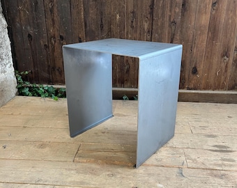 Könnte beinhalten: Kleiner, quadratischer Beistelltisch aus Metall mit hellgrauer Oberfläche. Der Tisch hat ein schlichtes, minimalistisches Design mit offenem Fuß. Er besteht aus einem einzigen Metallstück, das zu einer Kastenform gefaltet wurde.