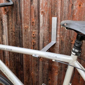 Op de afbeelding: Een zilveren fietsframe is met een metalen beugel aan een houten muur bevestigd. De fiets heeft een zwart zadel en stuur. De woorden "MAGIC TOUR" en "BIKE MANUFAKTU" zijn zichtbaar op het frame.