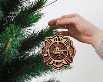May include: A wooden Christmas tree ornament in the shape of a fire department emblem. The ornament is brown and red with the words "FIRE DEPT." and "GODS SPEED" on it. The ornament is hanging on a green Christmas tree.