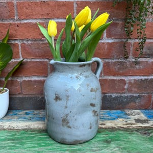 May include: A pale blue ceramic jug with a handle, holding bright yellow tulips and green leaves. The jug has a distressed, aged look. The background includes a red brick wall and a colourful wooden surface.