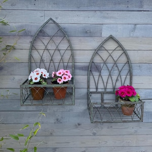 May include: Two metal wall planters with a gothic arch design. Each planter has a shelf and is filled with pink and white flowers.