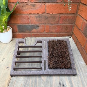 May include: A brown cast iron boot scraper with a built-in brush. The scraper has a rectangular shape with a decorative border and a section for scraping mud off shoes. The brush is made of stiff, brown bristles.