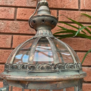 May include: A tall, ornate metal lantern with a glass dome and a decorative top. The lantern has a distressed, aged finish in shades of brown and green.