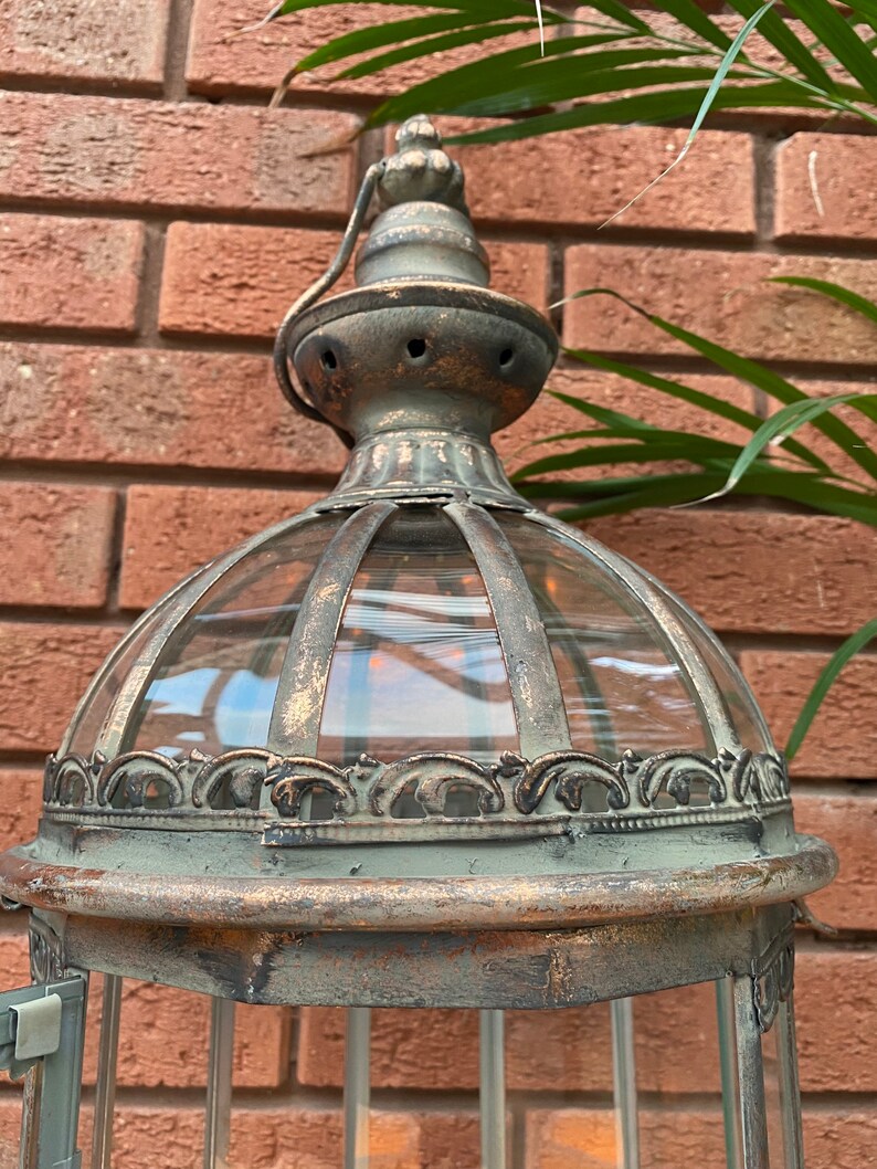 May include: A tall, ornate metal lantern with a glass dome and a decorative top. The lantern has a distressed, aged finish in shades of brown and green.