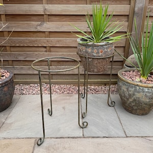 May include: Two metal plant stands with a decorative scroll design. The stands are painted a dark green colour and have a round top. The stand on the left is empty, while the stand on the right holds a potted plant with green leaves.