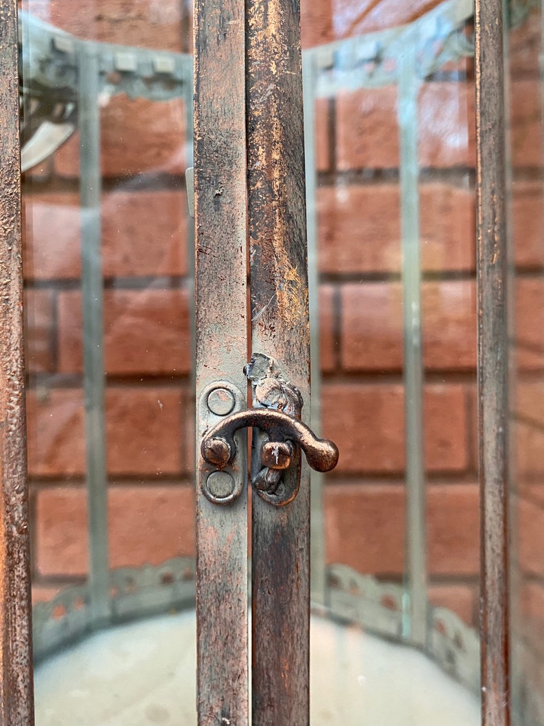 May include: A rustic metal lantern with a glass panel and a metal latch. The lantern has a distressed brown finish and a decorative design around the glass.