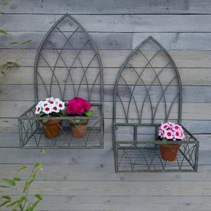 May include: Two metal wall planters with arched tops and a geometric design. The planters have shelves to hold potted plants. The planters are painted a weathered green colour. The planters are mounted on a wooden wall.