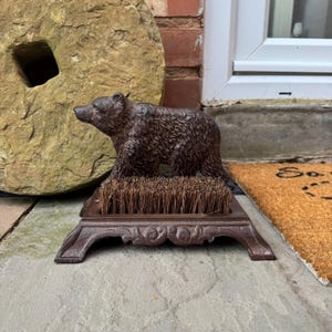 May include: A brown cast iron boot scraper shaped like a bear with a brown bristle brush. The scraper is on a stone surface.