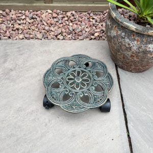 May include: An ornate, cast metal plant caddy with a floral design and a weathered green finish. The caddy has four black wheels for easy movement of potted plants. The caddy is designed to hold a large plant pot.