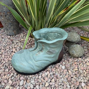 May include: A teal-coloured boot-shaped planter with a lace-up design and brown heel. The planter, made to resemble a worn boot, sits on a bed of small rocks. Green plants are visible in the background, adding a natural touch.